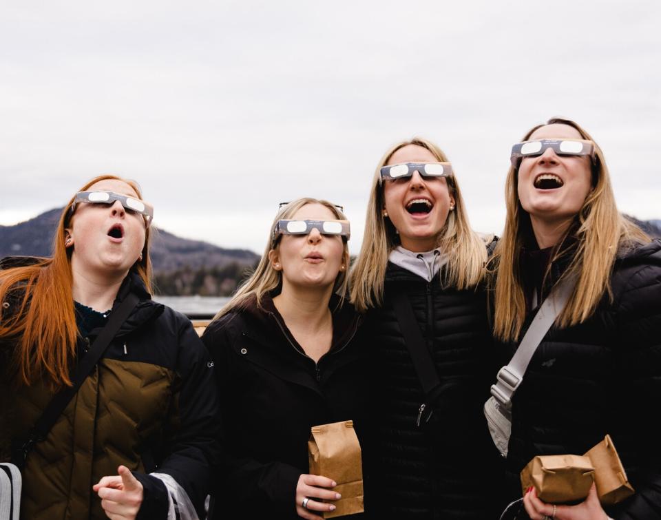 Three women wearing eclipse glasses look up into the sky in awe.