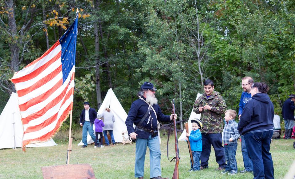 A man in a Union soldier uniform carrying a musket and talking with people attending the event. An American flag of that era is flying on a pole and there are white canvas tents in the background