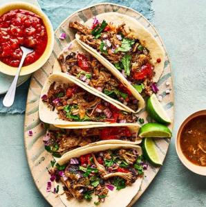 5 tacos placed on a plate in a fan-like display with lime wedges and a bowl of salsa on the side with a spoon it