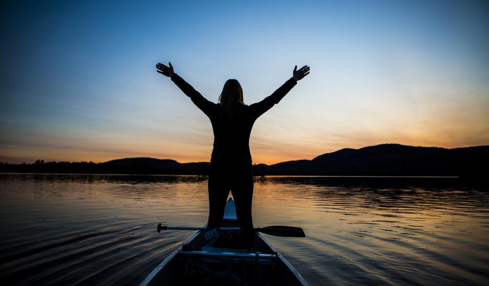A woman stands up with her arms up in a canoe.