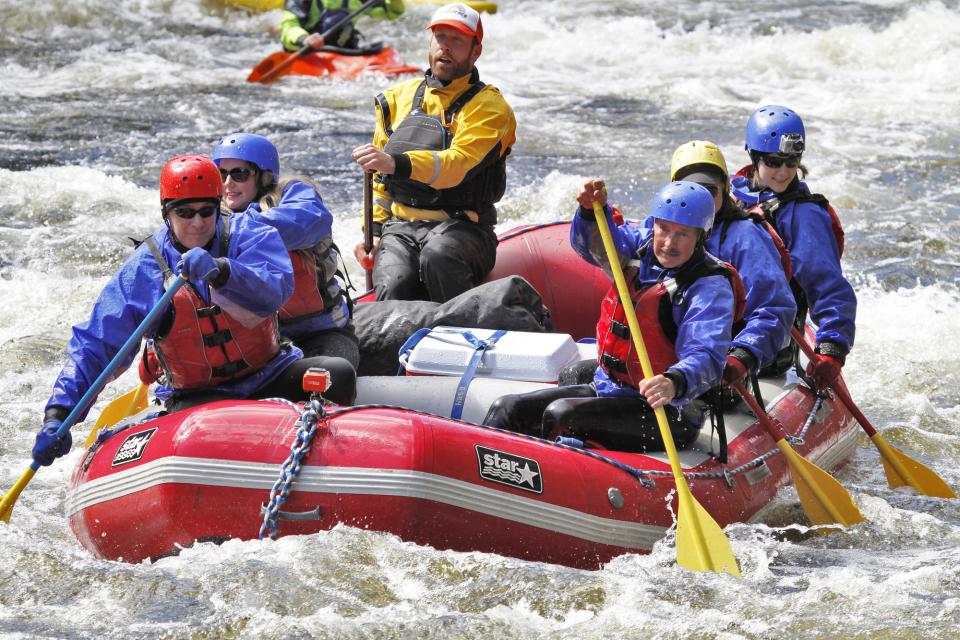 A group of people in PFDs and helmets paddle a raft on a churning river.
