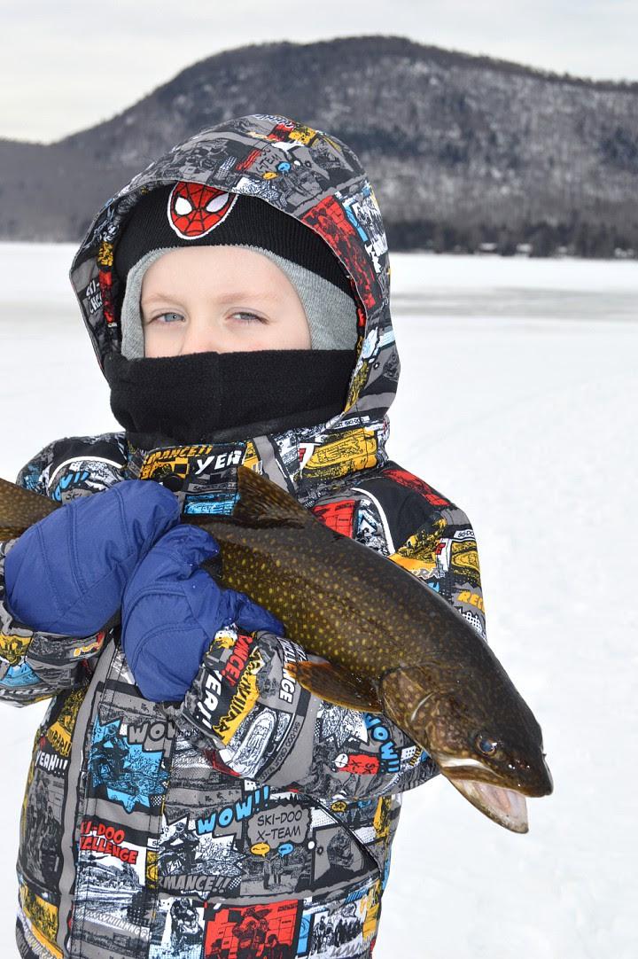 A young child in a colorful jacket holds a fish caught while ice fishing.