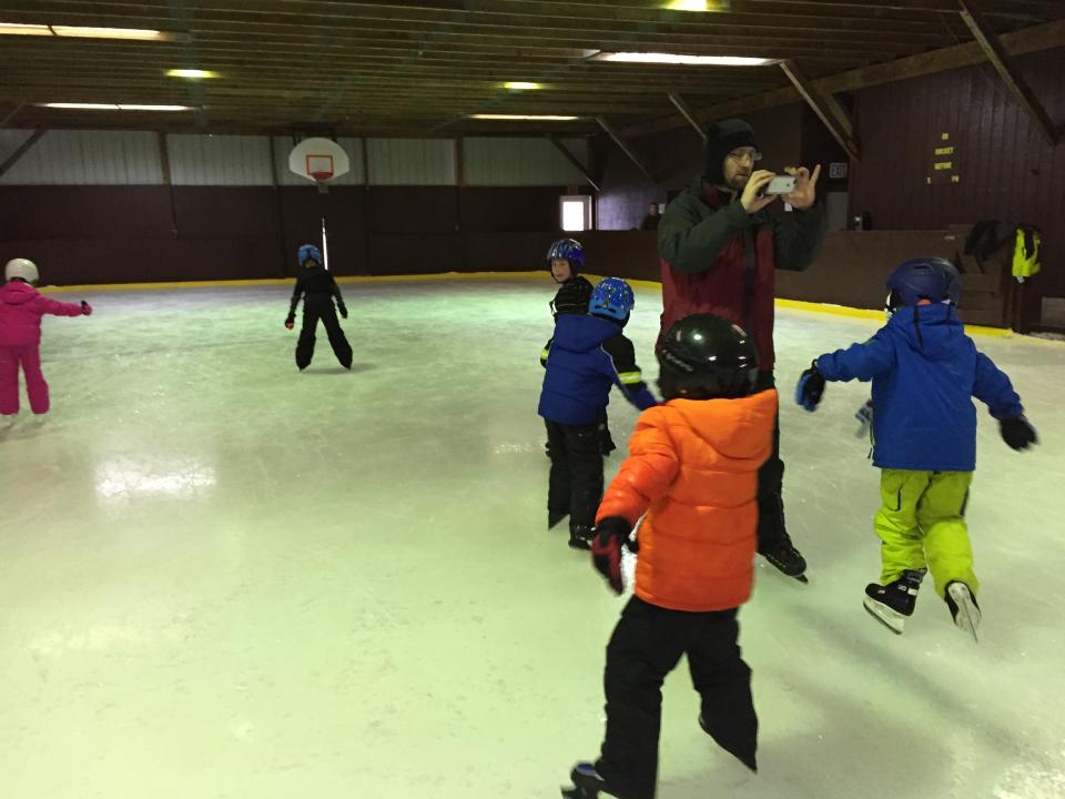 Children at an inclosed ice skating rink with a man taking their photo on a cell phone.