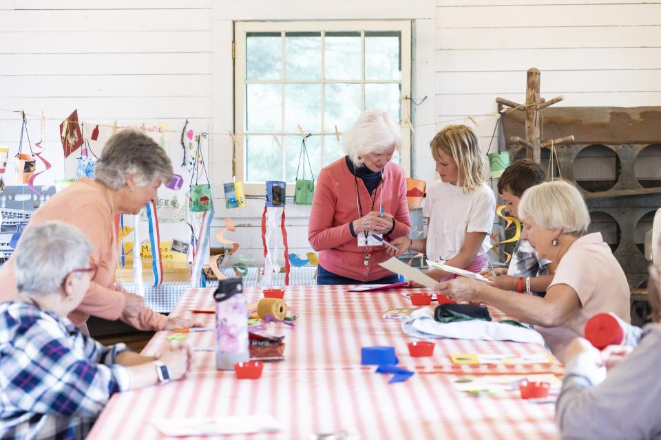 A brightly lit room is filled with smiling grandparents and grandchildren making colorful crafts.