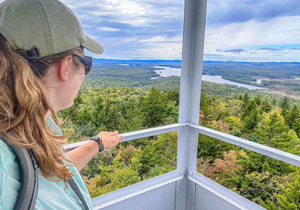 A hiker with a blonde ponytail and sunglasses look at a view of a lake from a fire tower.