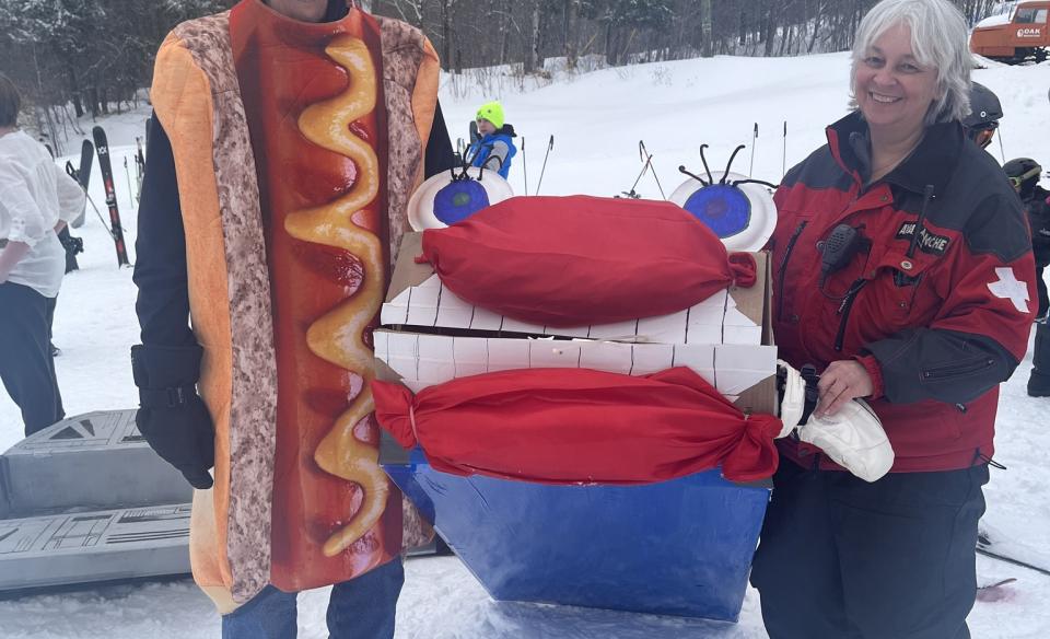 Person dressed up in a hotdog costume standing with a woman holding a cardboard sled between them that has a big goofy face on it.
