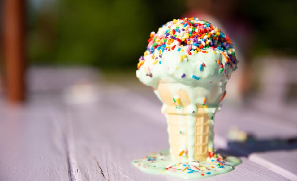 Ice cream cone with green ice cream and rainbow sprinkles standing on a picnic table with melting drips running down the side