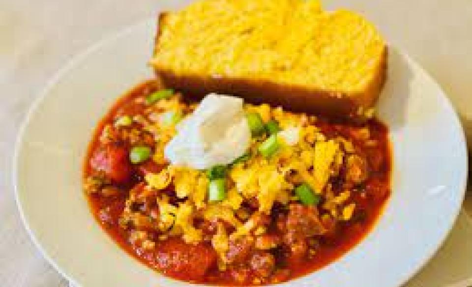 A bowl of chili with shredded cheddar cheese and sour cream on top with a side of corn bread
