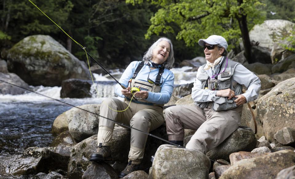 Two older women sitting on rocks on the edge of a river fly fishing and laughing