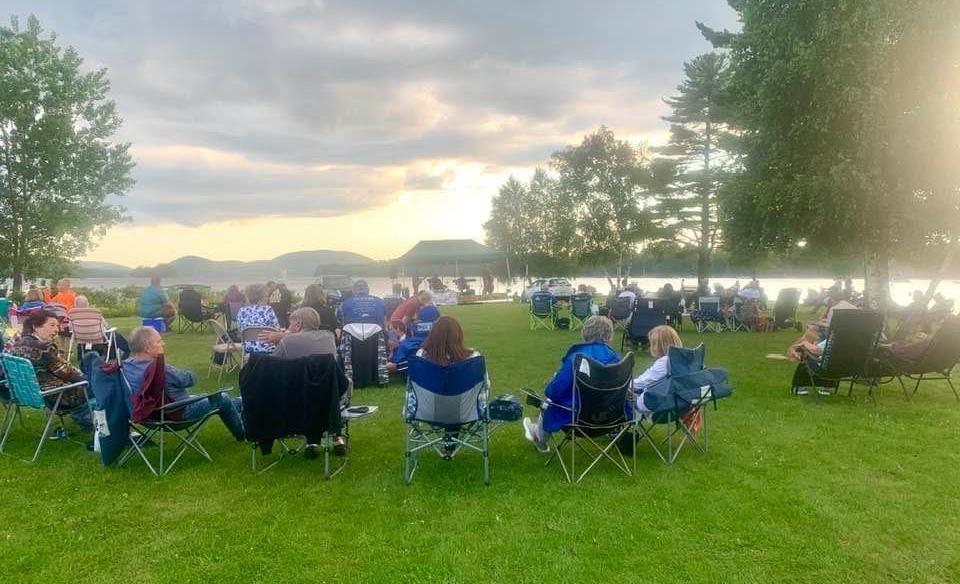 Groups of people sitting in camp chairs enjoying live music on the point at Speculator with Lake Pleasant in the background