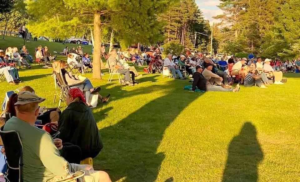 Lots of people sitting in camp chairs and on the ground listening to live music on the point in Speculator with the sun shining on them as it sets