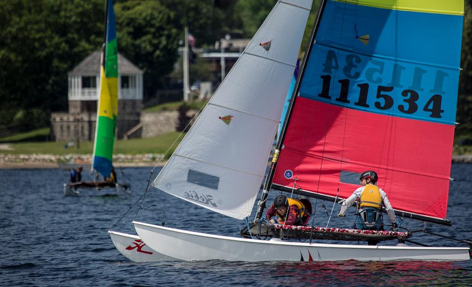 A sailboat with a white, red, blue, and green sail, on the lake with two people sailing it. Another sailboat is in the background with a green and yellow sail.