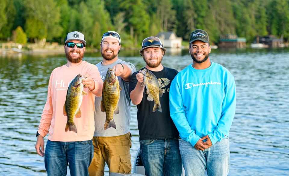 four men standing in front of a lake, three of them are holding up fish that they caught. All of them are smiling