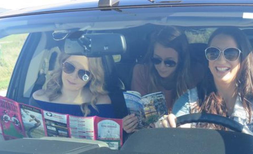 Three young women in a car, one is driving and the other two are looking at maps