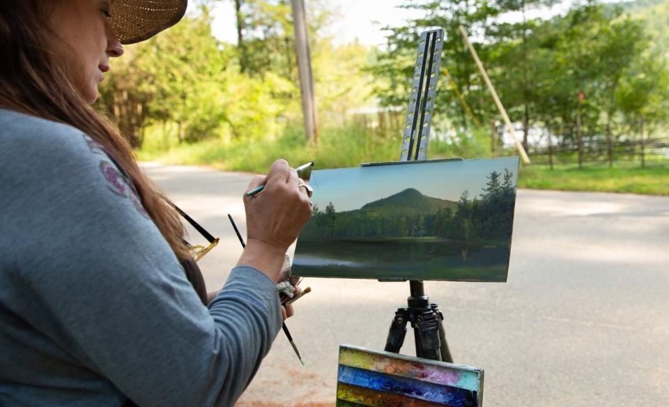 A woman in a blue long sleeve shirt and sun hat is out side painting the landscape that is before her, which include a lake with mountains in the background and a forest.