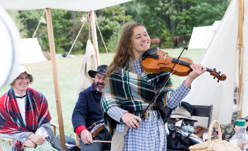 A young woman is playing a fiddle dressed in the Civil War era with others dressed in period clothing sitting in the background