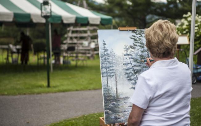 A woman stands with her back to the camera, painting a canvas en plein air.