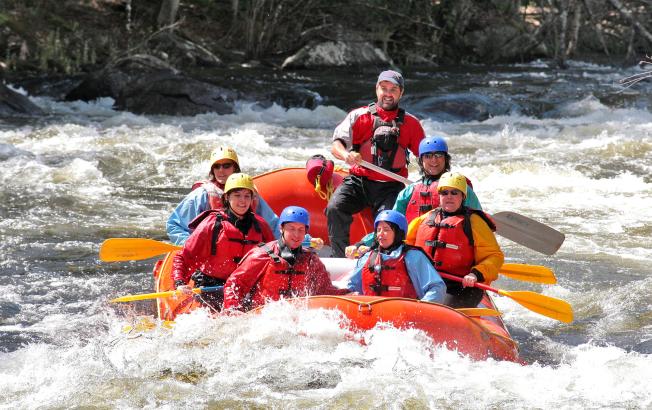 A group of adults wearing raincoats, life vests, and helmets paddle a raft on a roaring river.