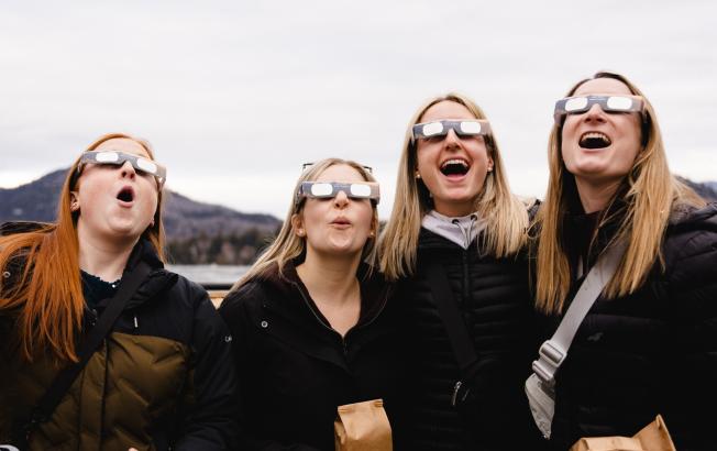 A group of four woman look to the sky with eclipse glasses