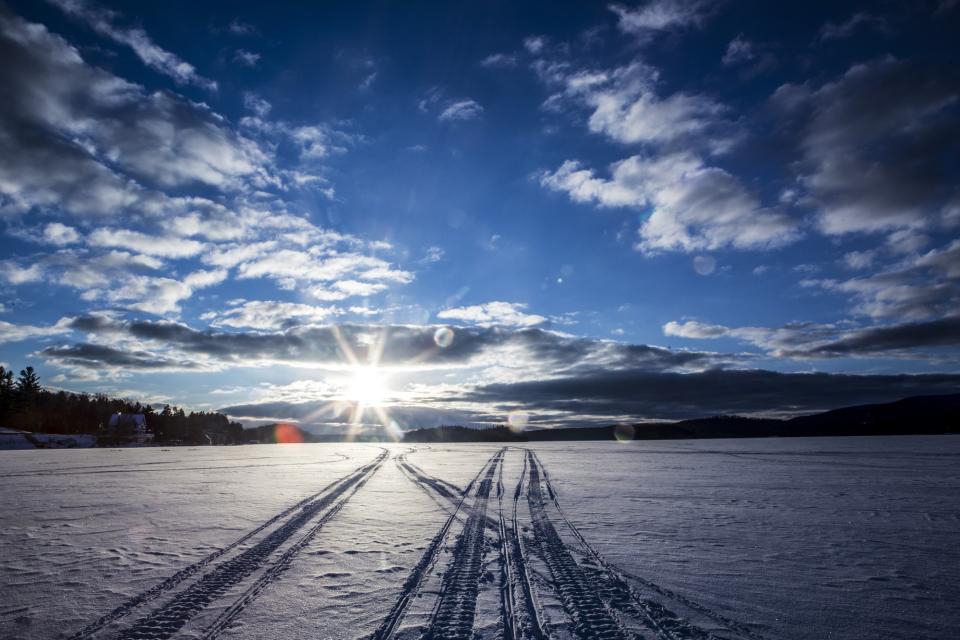 Snowmobile tracks extend to the horizon on a snowy, frozen lake under a bright blue sky.