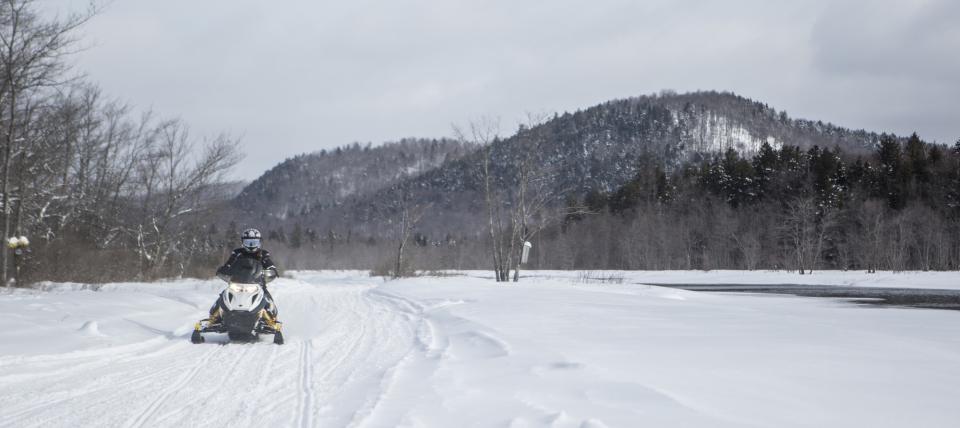 A rider on a snowmobile passes a snowy landscape and mountain.