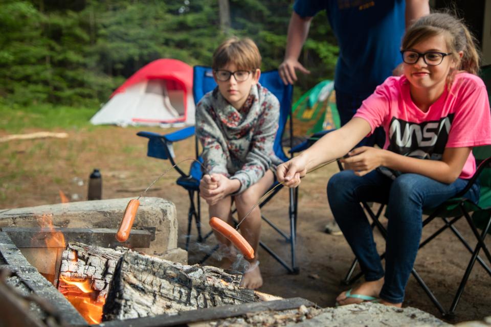 Two children roast hot dogs over a daytime campfire.