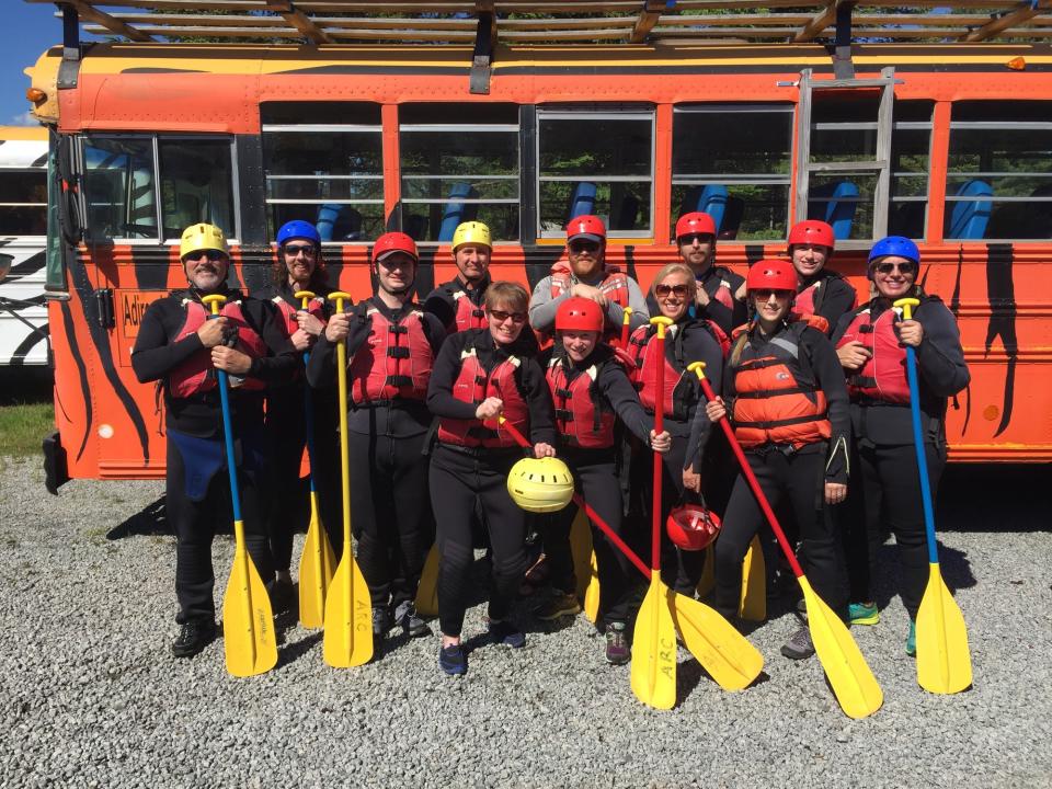A group of whitewater rafters pose with paddles in rafting gear in front of a bus.