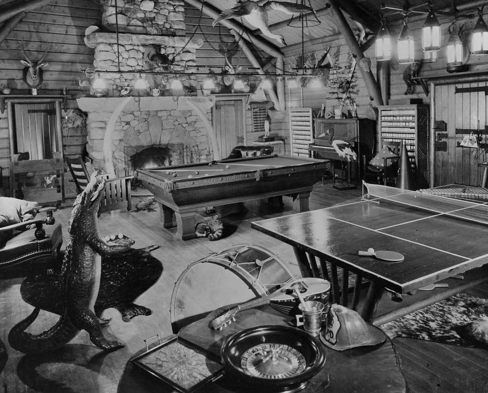 Adirondack Museum Collection. Photo description: Scene of game room at Great Camp Sagamore. Pool and ping-pong tables at center of room, various musical instruments around. Large fireplace at back of room. Mounted animals and birds throughout the room. Stuffed Aligator holding ping-pong balls at left, sleeping raccoon under pool table. Mounted to board with P6892 on reverse.