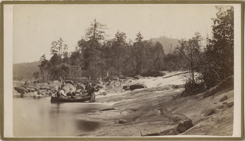 Photo print showing a group of men and women sitting in boats where Bog River Falls meets Tupper Lake, Catalog Number  1975.020.0697