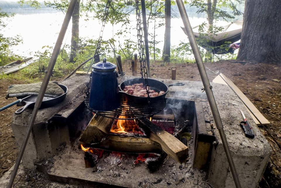 A pot of coffee and pan of bacon cooking in a pot over a campsite fire pit.
