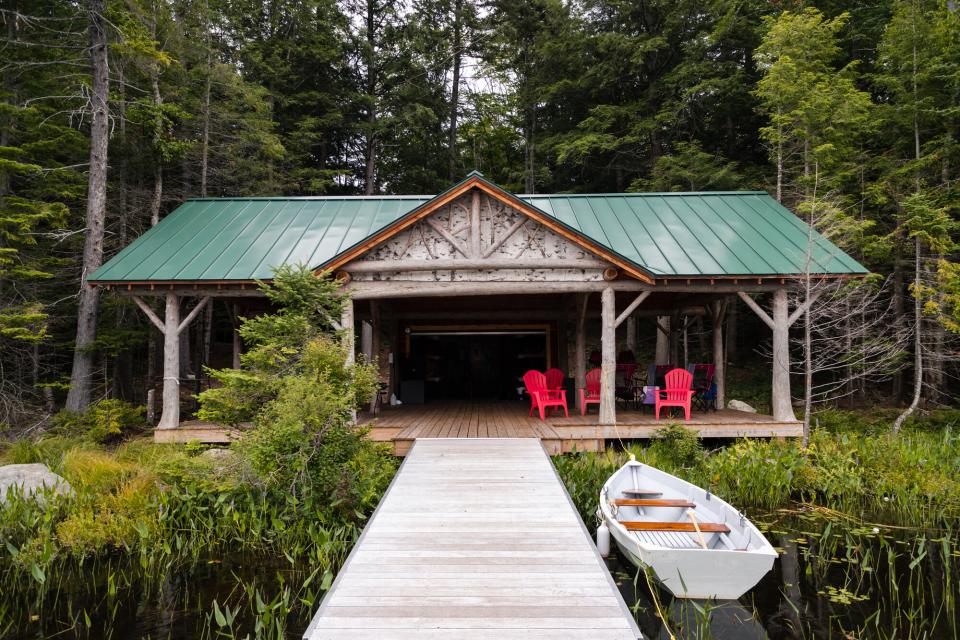 A rustic, bark-trimmed Adirondack boathouse with a long dock and a rowboat in the foreground.