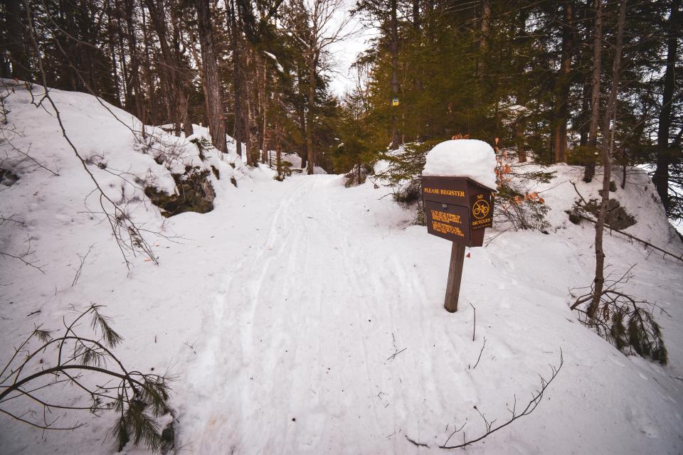 The sign-in register to the right of the trail with a large pile of snow on top.