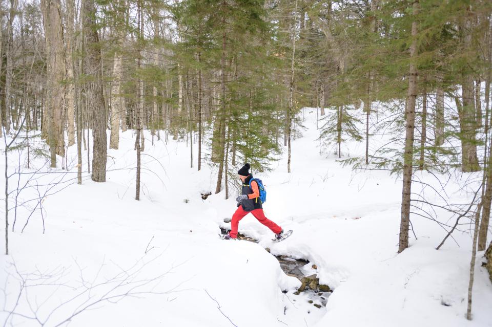 A woman in snow pants, coat, and winter hat crosses a small stream on snowshoes.