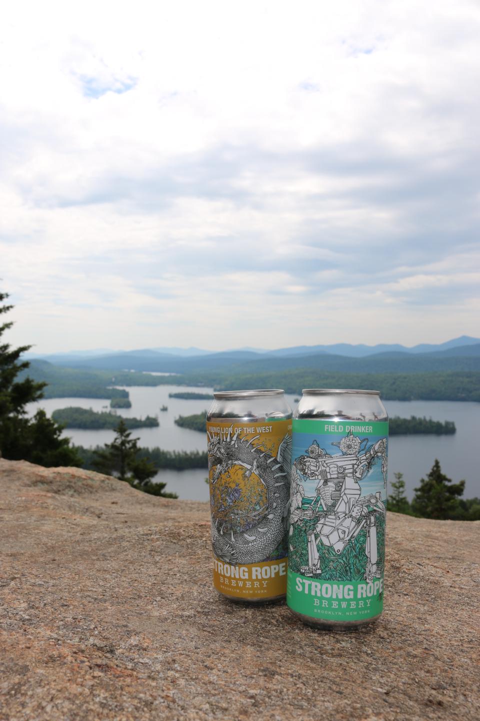 Close up of two cans of Strong Rope beer on a rock ledge with a lake in the background.