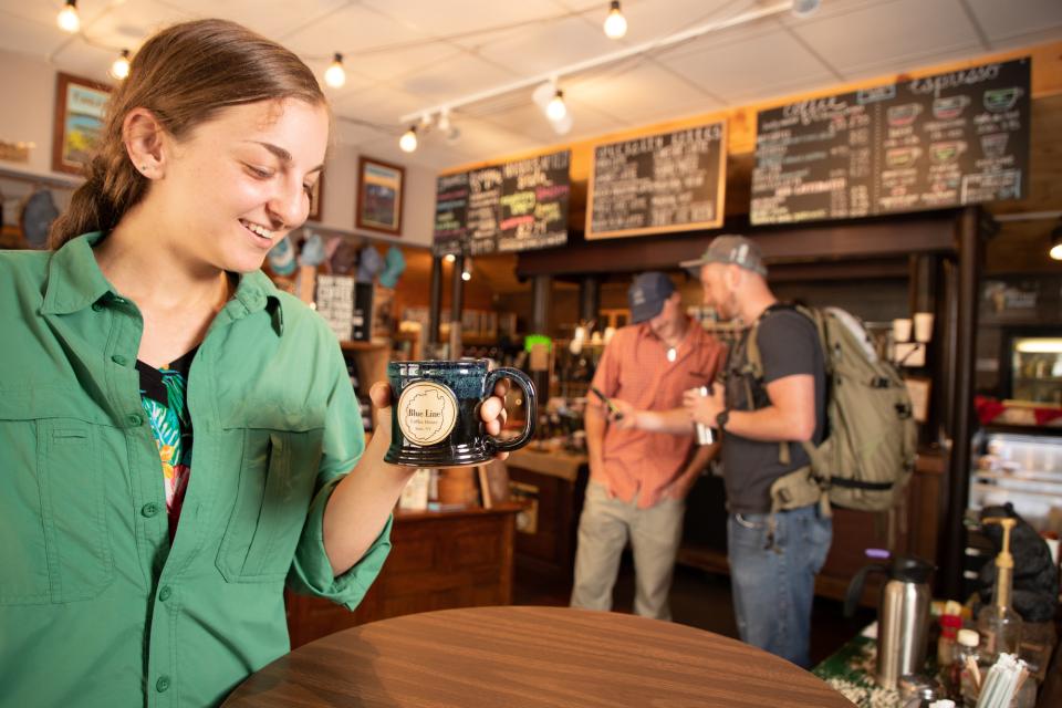 A woman smiles with a mug of coffee in a coffee shop while two men look at a cell phone behind her.
