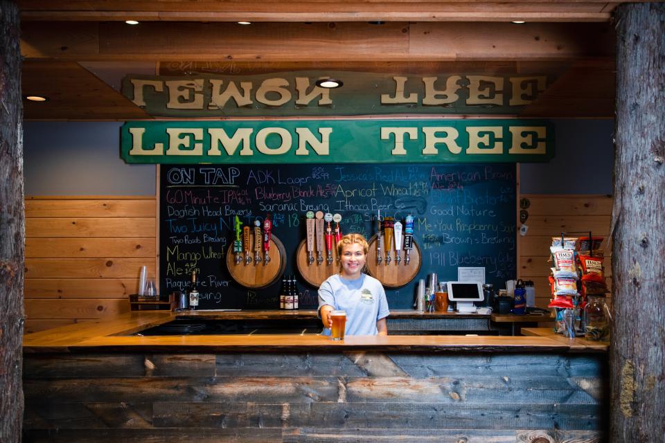 A smiling bartender holds out a glass of beer from behind a rustic wood bar with chalkboard menu behind her.