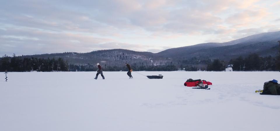 A couple pulls a sled across a snowy, frozen lake with mountains in the background.