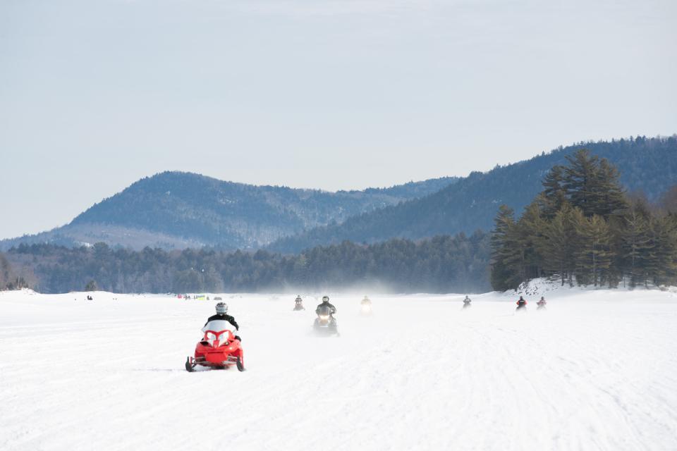 A group of snowmobiles zoom across a frozen lake with forested mountains in the background.