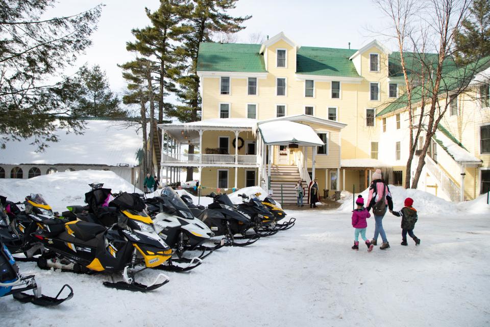 A person with small children walk past a row of snowmobiles parked in front of a large, old fashioned inn.