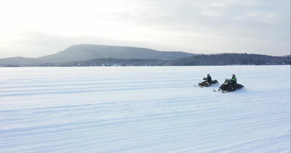 Two people ride their snowmobiles on a frozen lake with mountains in the background