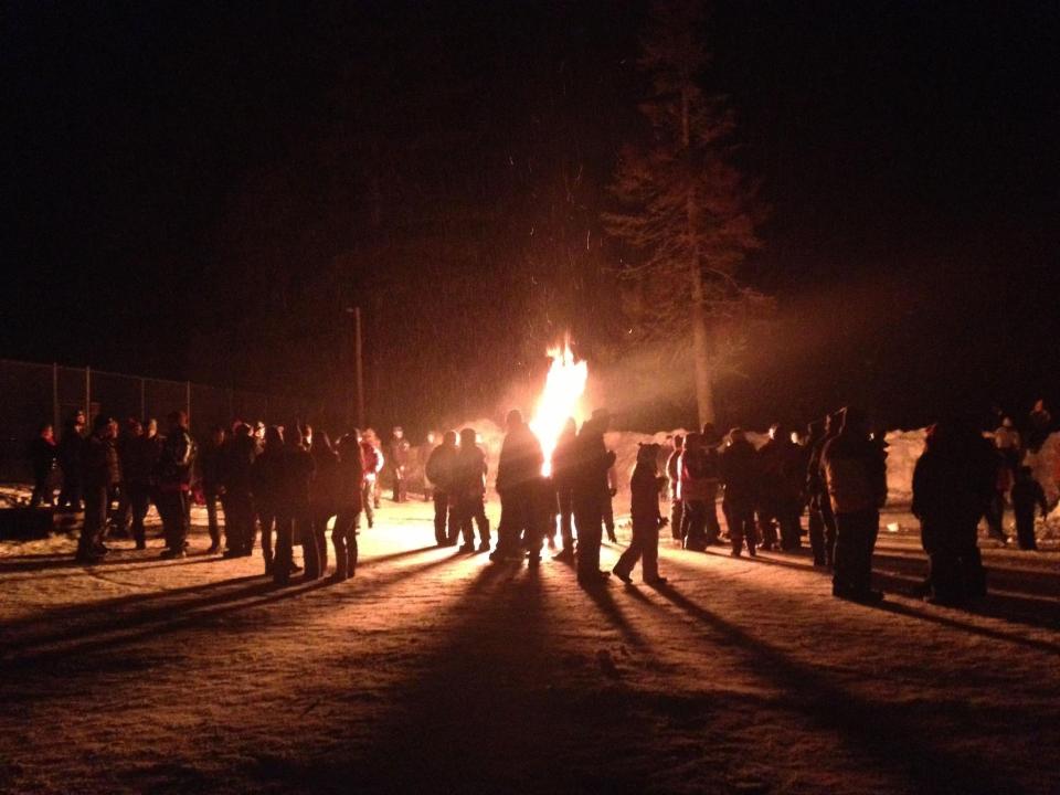 A large group of people stands around a large bonfire in winter.