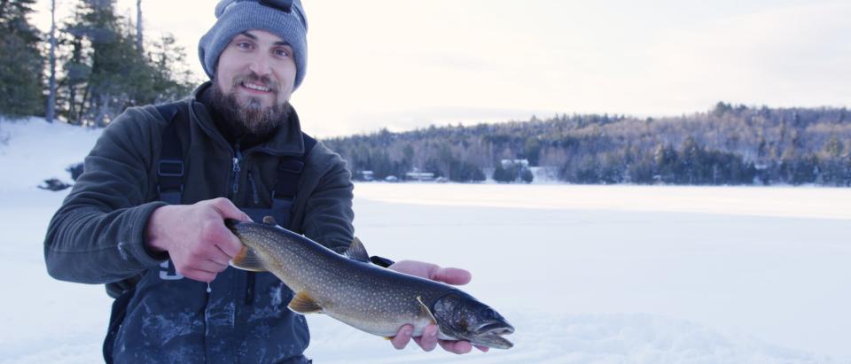 A man with a beard smiles as he holds a lake trout caught while ice fishing.