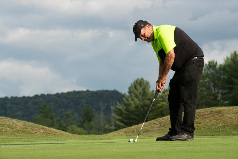 A golfer putts on a scenic green with trees and low hills in the background.