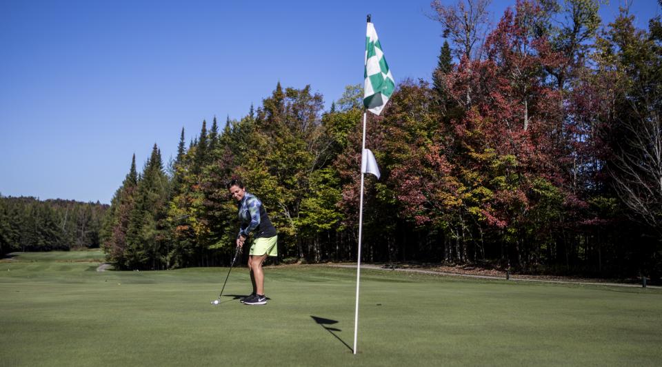 A woman putts on a golf green with fall-tinged trees behind her.