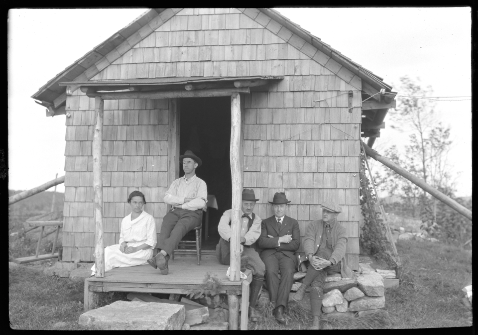 A black and white image of five people sitting outside an old cabin.
