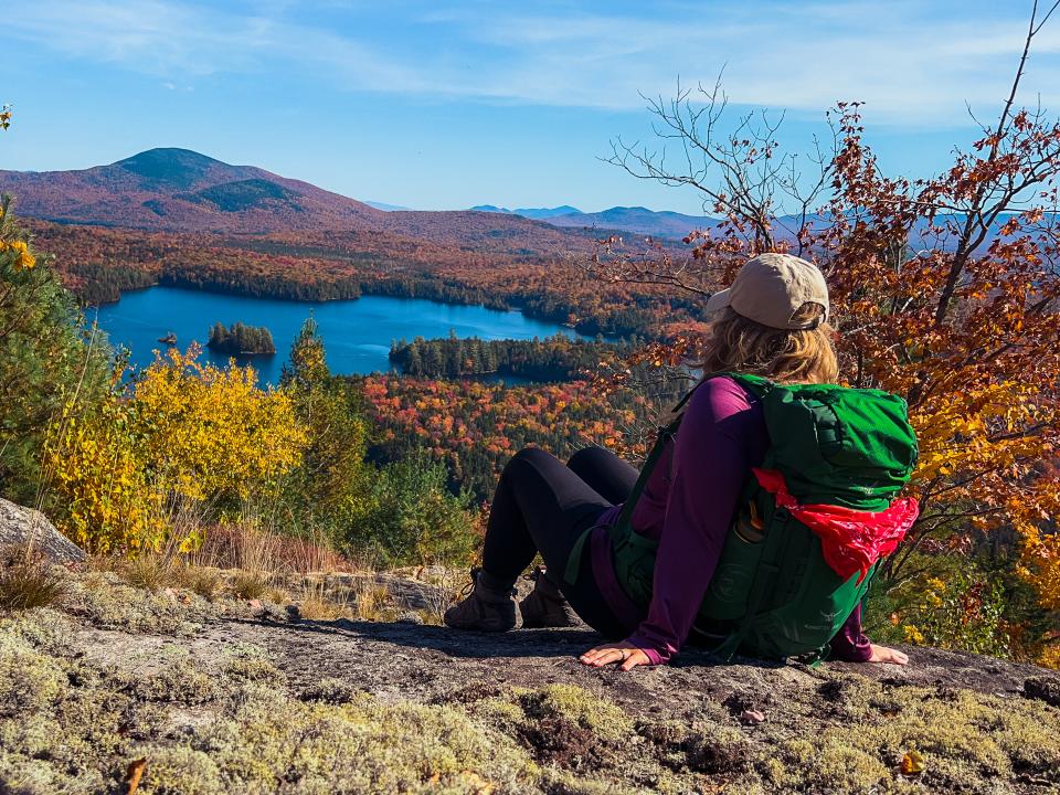 A woman sits on a mountain summit looking out at a pond and forest vibrant with orange and yellow leaves.