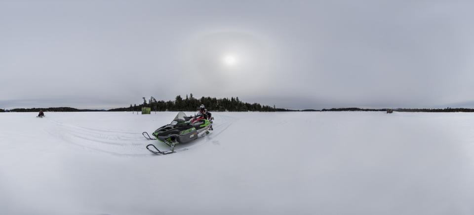 A snowmobiler drives a snowmobile across a large frozen Adirondack lake with a fishing ice shanty in the distance.