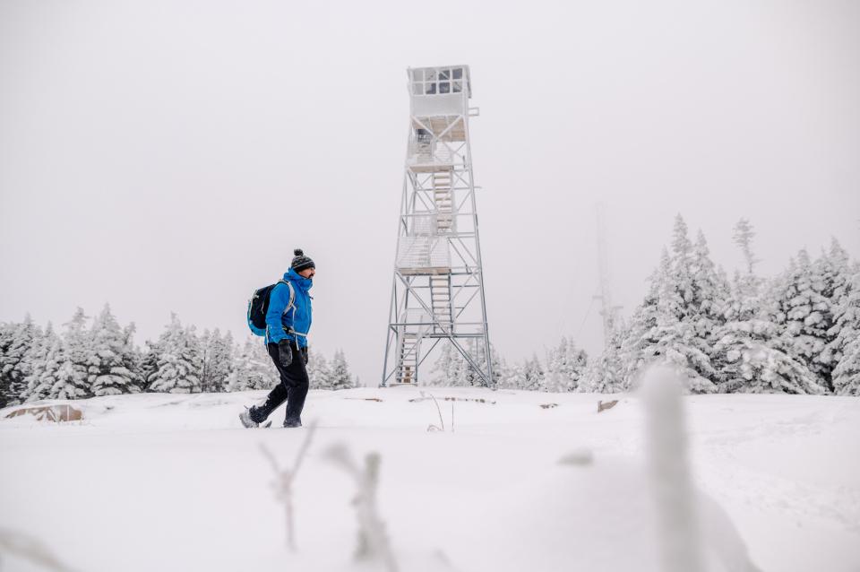 A snowshoer in a bright blue coat walks on a summit near a fire tower; the whole landscape is white with snow