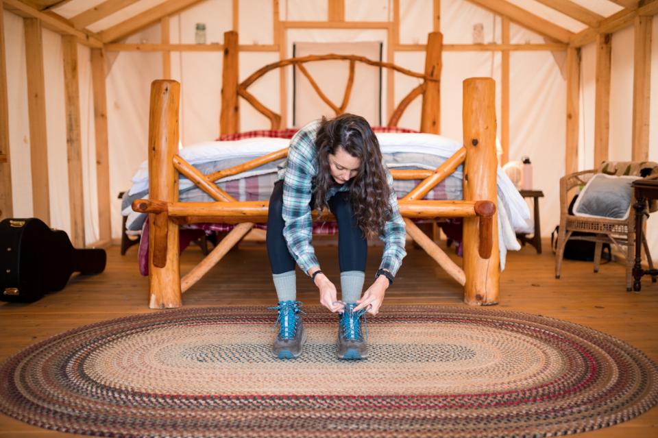 A woman ties her hiking boots at the foot of a rustic bed.