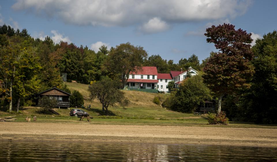 A white, farmhouse-style inn on a small hill with sandy beach in front. Photo taken from the water.